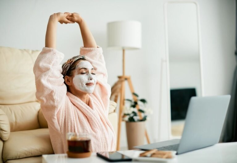 On international self care day, a person wearing a face mask and bathrobe stretches with eyes closed while sitting in front of a laptop, with tea and snacks on the table.