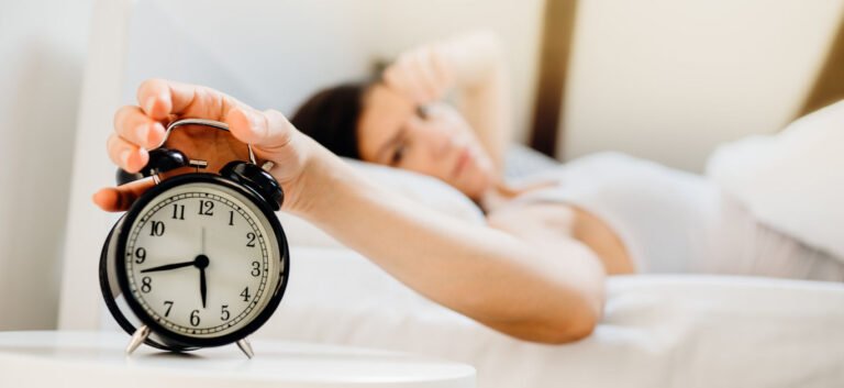How stress physically changes your sleep patterns Person lying in bed reaching to turn off a black analog alarm clock on a white nightstand at wake-up time