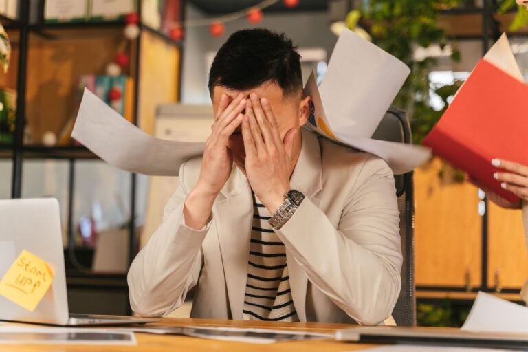 Financial stress is a direct health risk Stressed businessman overwhelmed by paperwork in office environment, demonstrating burnout.