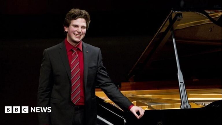 British-Australian pianist Jayson Gillham wearing a suit stands beside a grand piano.