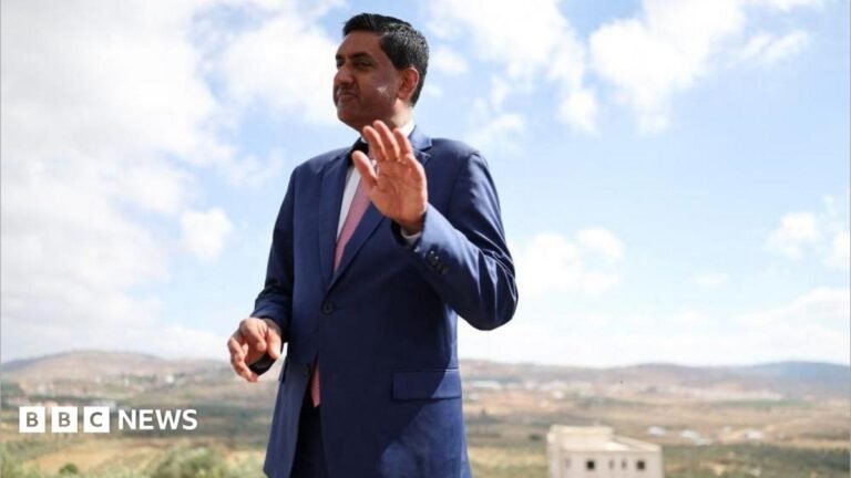 US Democrat Ro Khanna says he was detained by armed Israeli settlers Khanna, who is dressed in a blue suit, white shirt and pink tie, raises his hand as he is photographed outdoors with a rural town landscape behind him