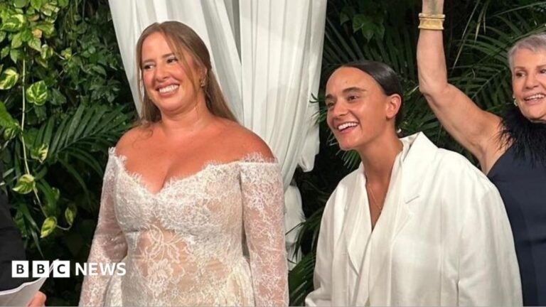 Former British-Israeli Hamas hostage married in Tel Aviv Two women hold hands in white bridalwear; one wears a white tailored suit, while the other wears a lace dress.