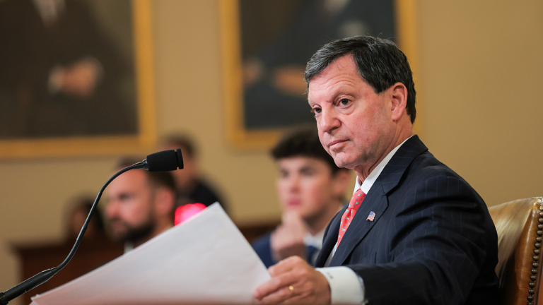 Social Security’s Countdown Clock is Getting Louder A man in a suit holds papers and sits at a desk with a microphone, looking to the side. Two blurred people and framed portraits are visible in the background.