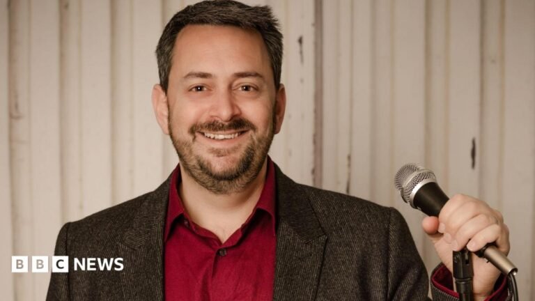 Venue admits scrapping Jewish comedian’s Edinburgh Fringe show was discrimination A dark-haired man who is wearing a dark grey blazer over a red shirt, holding a microphone stand and smiling.
