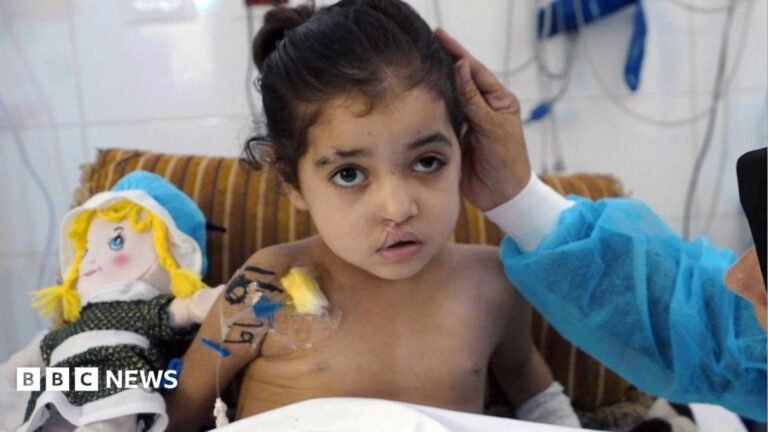 A young girl is seen in a hospital bed while a woman wearing scrubs strokes her hair