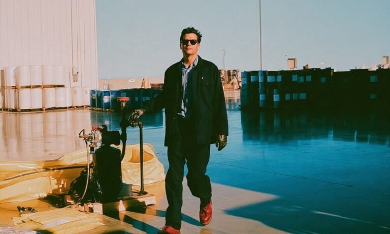 a successful plant manager in a tailored work jacket walking a fluid transfer line with his gloved hand resting near a pump and check valve assembly in his business warehouse