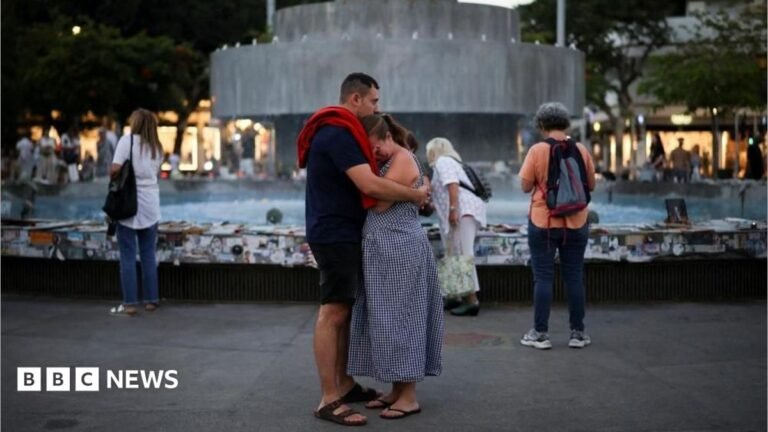 Israelis mourn 7 October attack victims as Gaza lies in ruins three years on A man and woman embrace in front of the fountain at Dizengoff Square. The woman has her hand to her eyes as if she is crying.